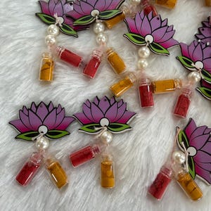 May include: Close-up of multiple decorative lotus flower ornaments. Each ornament features a purple and pink lotus flower design with green accents, connected to small glass vials filled with red and yellow powders. The ornaments are strung with faux pearls.