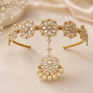 May include: A gold-toned metal headband featuring five circular floral motifs set with clear, teardrop-shaped stones and bordered by small white pearls. A central dangling pendant with matching stones and a row of hanging cream-colored pearls.