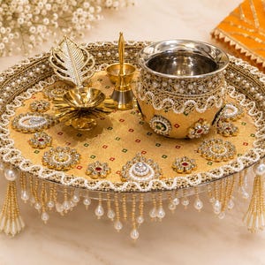 May include: A round gold decorative tray featuring intricate beadwork, pearl-like accents, and hanging beaded fringe. The tray holds a metallic pot with ornate embellishments, a gold-toned leaf-shaped object, and a small brass-colored bell.