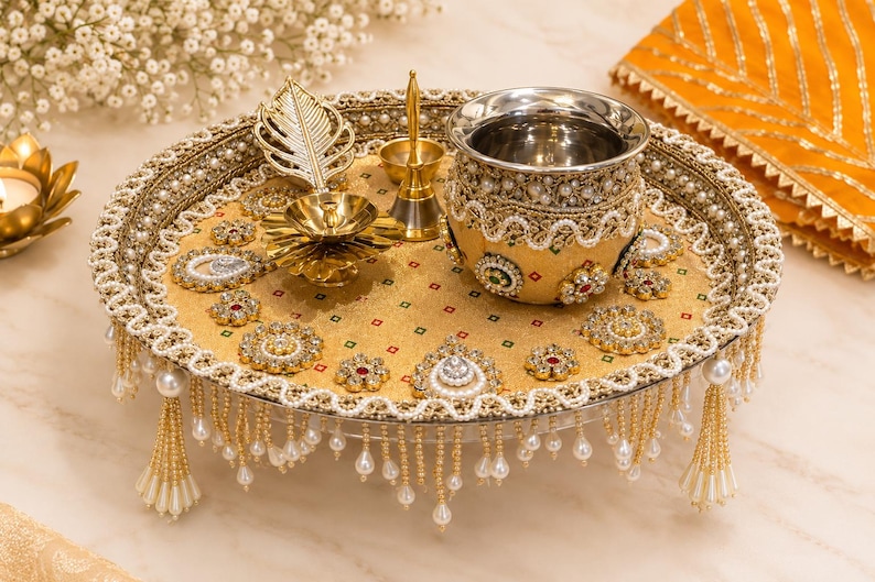 May include: A round gold decorative tray featuring intricate beadwork, pearl-like accents, and hanging beaded fringe. The tray holds a metallic pot with ornate embellishments, a gold-toned leaf-shaped object, and a small brass-colored bell.