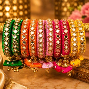 May include: A collection of colorful bangles in shades of green, orange, pink, and yellow. Each bangle is adorned with mirrored accents and decorative tassels. The bangles are displayed on a gold rod, showcasing their intricate design and vibrant colors.