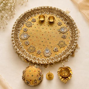 May include: A circular gold-toned decorative tray featuring a textured fabric surface with small multi-colored square patterns, embellished with ornate floral-shaped metal accents, clear teardrop-shaped stones, and a wide, intricate lace border.