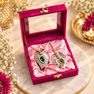 May include: A magenta velvet jewelry box with a clear lid containing two silver-toned brooches. One brooch is a teardrop shape with a central green stone and clear accents. The second brooch is a feather design with a small peacock eye detail.