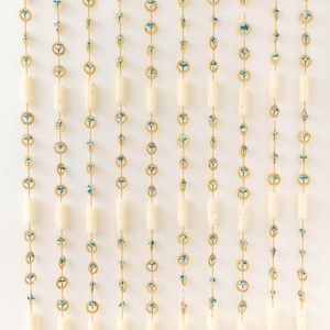May include: Decorative hanging garlands with cream-colored beads, gold accents, and blue floral designs. The garlands are suspended from white hooks and terminate in gold bell-shaped ornaments, creating an elegant, vertical display.
