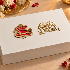 May include: A rectangular white gift box featuring a gold-toned Ganesha emblem with a red inlay and the gold-toned script text Happy Diwali centered on the lid. The box has a smooth, matte finish and a clean, minimalist design.