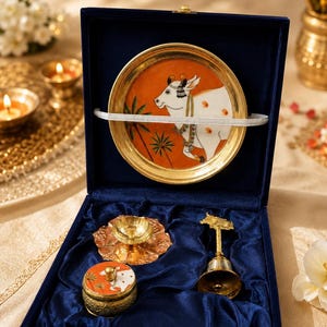 May include: A navy blue velvet gift box containing a brass plate with a painted white cow, a small brass oil lamp, a matching brass lidded container with a painted cow, and a brass bell with a cow figure on top, all set in blue fabric.