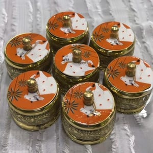 May include: A set of six round brass containers with lids. Each lid features an orange and white enamel design depicting a stylized white bull with gold accents and a small gold-toned knob handle. The brass sides have an intricate embossed pattern.
