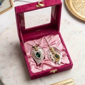 May include: A velvet jewelry box in a deep pink hue, open to reveal a silver and gemstone pendant and a feather-shaped accessory. The box is lined with pink satin. A gold nameplate reads "Roli Chopra's Creations."