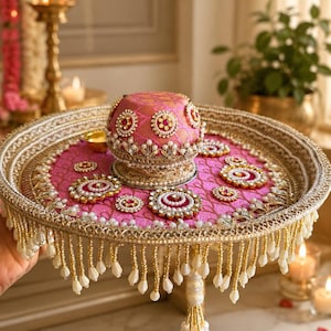 May include: A round decorative tray featuring a vibrant pink fabric base with gold embroidery, adorned with circular floral-shaped embellishments. A matching pink dome-shaped cover sits in the center, surrounded by hanging white and gold beaded fringe.