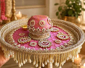 May include: A round decorative tray featuring a vibrant pink fabric base with gold embroidery, adorned with circular floral-shaped embellishments. A matching pink dome-shaped cover sits in the center, surrounded by hanging white and gold beaded fringe.