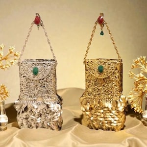 May include: Two rectangular handbags, one silver-toned and one gold-toned, featuring intricate filigree metalwork, a central green oval stone on the flap, a chain handle with a red accent, and a base covered in overlapping metallic circular discs.