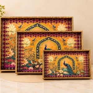 May include: A set of three rectangular nesting trays with gold-toned frames. Each tray features a vibrant orange background, a central archway, a peacock, lotus flowers, and small deer, all bordered by a deep magenta pattern with gold accents.