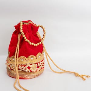 May include: A red velvet drawstring bag with gold beaded handle and drawstring. The bag is adorned with gold embroidery, pearls, and sequins along the bottom edge. A decorative and elegant accessory.