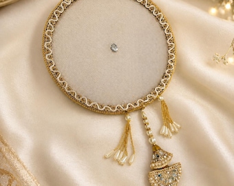 May include: A circular beige fabric ornament featuring a gold embroidered border, a single central crystal, and dangling gold tassels with small white beads. A decorative gold pendant with clear stones and pearls hangs from the bottom edge.