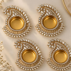 May include: Four gold-colored metal diya candle holders shaped like paisley motifs. Each holder features a central circular cup surrounded by a decorative border of small, clear, reflective mirror-like pieces and embedded white crystals.