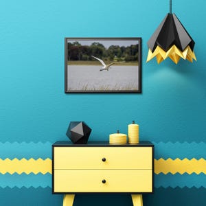 May include: A framed photograph of a white bird in flight over a body of water hangs on a teal wall. Below, a yellow dresser with black accents holds decorative items. A black and gold geometric pendant light hangs from the ceiling.