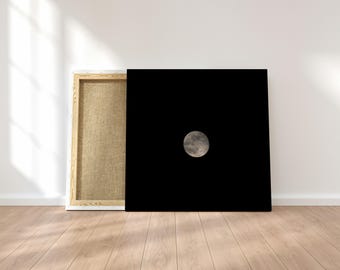 May include: A square canvas print featuring a solid black background with a single, centered, textured gray moon. The canvas is mounted on a wooden frame, showing the side profile of the stretcher bars and the raw, light brown fabric texture.