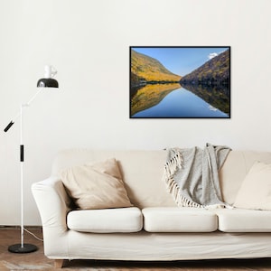 May include: A framed landscape photograph of a lake reflecting the surrounding mountains and sky. The mountains are covered in autumn foliage, with shades of yellow and orange. The sky is a clear blue, and the water is calm.
