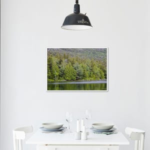 May include: A framed landscape photograph of a forest and lake. The image features a lush green forest bordering a calm body of water. The photograph is displayed on a white wall above a white dining table with place settings.