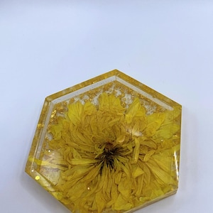 May include: A hexagonal coaster with a yellow flower encased in clear resin. The flower is centered, with gold glitter accents throughout the resin. The coaster is a decorative item.