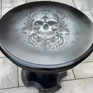 Peut inclure: Une table d'appoint ronde noire avec une illustration détaillée de crâne gris et blanc centrée sur le plateau. Le crâne est entouré de motifs floraux et de feuilles complexes. La table a une surface lisse et un pied central aux bords incurvés.