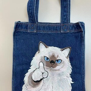 Puede incluir: Bolso tote de mezclilla azul con un diseño de gato bordado. El gato tiene ojos azules y está mostrando el dedo medio. El bolso tiene dos asas de mezclilla y es adecuado para transportar objetos.