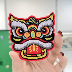 May include: Embroidered patch depicting a lion dance head. The patch is predominantly red, with yellow, white, and black detailing. The lion's face has large eyes, a white mustache, and an open mouth. The patch is suitable for clothing.
