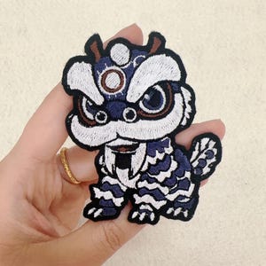 Op de afbeelding: Geborduurde patch met een gestileerde leeuwendansfiguur. De patch heeft een marineblauwe body met witte en bruine accenten. Het gezicht van de leeuw heeft grote, expressieve ogen en een gedetailleerde manen. De patch is waarschijnlijk bedoeld voor kleding of accessoires.