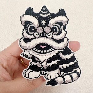 Puede incluir: Parche bordado en negro, blanco y gris con una figura estilizada de danza del león. El parche tiene un diseño detallado con una melena blanca prominente, un tocado decorativo y una boca sonriente. Para ropa o accesorios.
