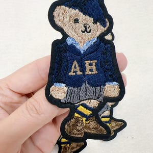 Puede incluir: Parche bordado con un oso de peluche con una blazer azul marino con letras doradas "AH". El oso lleva un gorro azul, una camisa azul claro y pantalones cortos grises. Tiene un borde negro y zapatos marrones con calcetines a rayas amarillas y azules.