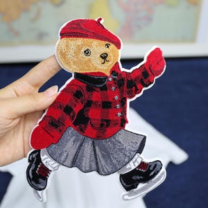 Może przedstawiać: Haftowana naszywka przedstawiająca misia w czerwono-czarnej kraciastej kurtce, berecie i łyżwach. Miś jest w pozie łyżwiarskiej, w szarej spódniczce i białych skarpetkach. Naszywka ma białą obwódkę.