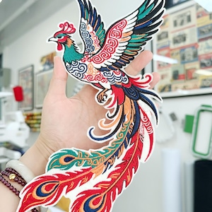 May include: A colorful embroidered patch featuring a phoenix with outstretched wings. The patch showcases a vibrant design with red, blue, green, and orange hues. The phoenix has a detailed pattern and is likely for decorative purposes.