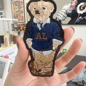 May include: Embroidered patch featuring a teddy bear wearing a navy blue sweater with the letters "AL" in brown. The bear is in a standing pose, with tan pants and a black border. The patch is held in a hand.