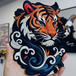 Puede incluir: Parche bordado con la cabeza de un tigre con detalles en naranja, negro y blanco. El tigre tiene ojos azules y está rodeado de olas estilizadas azules y blancas. Diseño detallado y colorido.