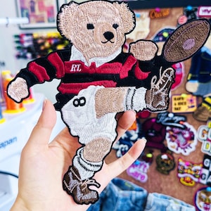 May include: Embroidered patch featuring a teddy bear in a rugby uniform. The bear wears a red and black striped jersey with "RL" on the chest and white shorts. It's holding a brown rugby ball and kicking a brown shoe. The patch is tan, black, red, and white.