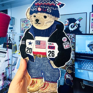 Puede incluir: Parche bordado de un oso de peluche con una chaqueta azul y negra, vaqueros azules y un gorro azul con la palabra "FAMOUS". La chaqueta presenta la bandera americana y los números "20" y "26". El oso lleva botas marrones.