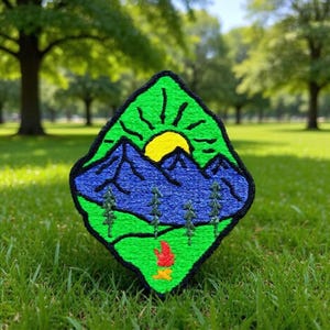 Puede incluir: Parche bordado con un paisaje de montaña con montañas azules, un sol amarillo y árboles verdes. El parche tiene un borde verde y una pequeña fogata en la parte inferior. El parche está sobre un fondo de hierba verde y árboles.