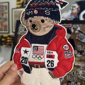May include: Embroidered patch featuring a teddy bear wearing a navy blue hat with "FAMOUS" text, a red jacket, and white pants. The patch includes the American flag, Olympic rings, and the numbers "20" and "26".