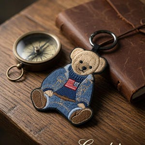 Op de afbeelding: Een geborduurde sleutelhanger van een beige teddybeer in een blauw spijkerjasje over een marineblauwe trui met een kleine Amerikaanse vlag. De beer heeft bruine pootjes en zit aan een zwarte metalen ring. De tekst Coretrims is zichtbaar.