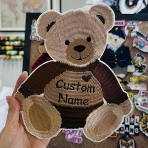 Può includere: Toppa ricamata a forma di orsetto nei toni del marrone, beige e nero. L'orso ha un cuore e le parole "Custom Name" ricamate sul petto. La toppa è tenuta in mano, con altre toppe e forniture per cucire sullo sfondo.