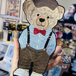 Puede incluir: Parche bordado de un oso de peluche con gorra marrón, camisa azul claro con tirantes, pajarita roja y pantalón marrón. El oso tiene cara color canela y zapatos blancos. El parche es probablemente para ropa o accesorios.