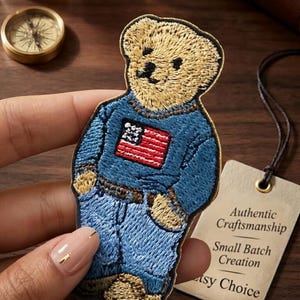 May include: Embroidered patch of a teddy bear wearing a blue sweater and jeans, with an American flag on the sweater. The patch has the text "Premium Embroidery • Vintage Vibes" at the top. A tag reads "Authentic Craftsmanship, Small Batch Creation, Etsy Choice."