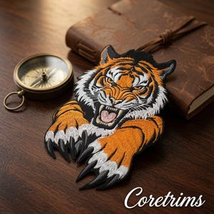May include: An embroidered patch featuring a snarling tiger head with orange, black, and white fur, baring its teeth and extending two large paws with sharp black claws. The patch is positioned near a vintage-style compass and a leather journal.