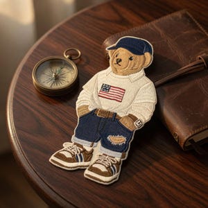 Op de afbeelding: Geborduurde patch van een teddybeer met een witte trui met een Amerikaanse vlag, een blauwe spijkerbroek en een marineblauwe pet. De beer draagt ook bruine en witte sneakers. De patch ligt op een donkere houten ondergrond.
