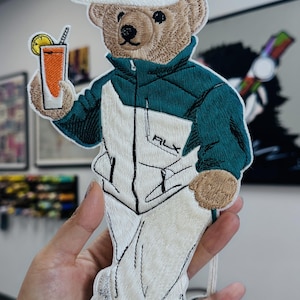 Puede incluir: Parche bordado de un oso con gorra blanca con una "A" verde y una chaqueta blanca y verde azulado. El oso sostiene una bebida con una rodaja de limón. El parche tiene las letras "RLX" en la chaqueta.