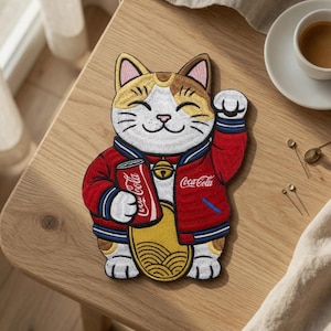 Könnte beinhalten: Ein bestickter Aufnäher, der eine Glückskatze in einer roten Jacke mit blau-weiß gestreiften Bündchen zeigt. Die Katze hält eine rote Coca-Cola-Dose in einer Pfote und hat ein goldenes Oval auf dem Bauch sowie ein Halsband mit einer goldenen Glocke.