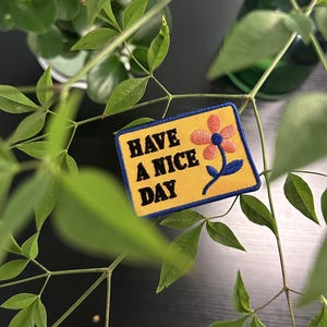Op de afbeelding: Een gele patch met de tekst "HAVE A NICE DAY" in het zwart. De patch heeft een oranje en blauwe bloem. De patch heeft een blauwe rand en is geplaatst tegen een achtergrond van groene bladeren en een donkere ondergrond.