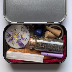May include: A metal tin containing a collection of items. Inside are a small glass bottle with a cork, a white candle with purple petals, a selenite wand, a pentagram charm, a blue gemstone, and other small objects. The tin is on a white surface.