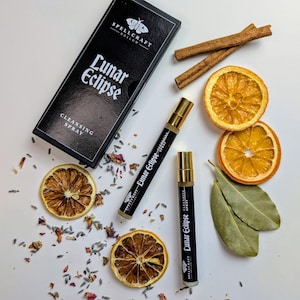 May include: Two spray bottles and a black box, all labelled "Lunar Eclipse Cleansing Spray". The image features dried orange slices, cinnamon sticks, and herbs, suggesting a natural product. The bottles have gold accents.