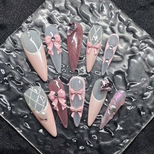 Könnte beinhalten: Eine Sammlung von zehn langen, mandelförmigen Kunstnägeln. Die Nägel weisen eine Vielzahl von Designs auf, darunter rosa und graue Farbverläufe, Schleifen, Perlen und dekorative Verzierungen. Einige Nägel haben ein klares, glänzendes Finish.